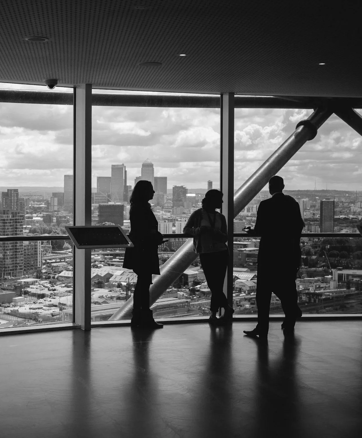 Silhouettes of three people talking inside a high-rise building with a city skyline view through large windows in black and white.