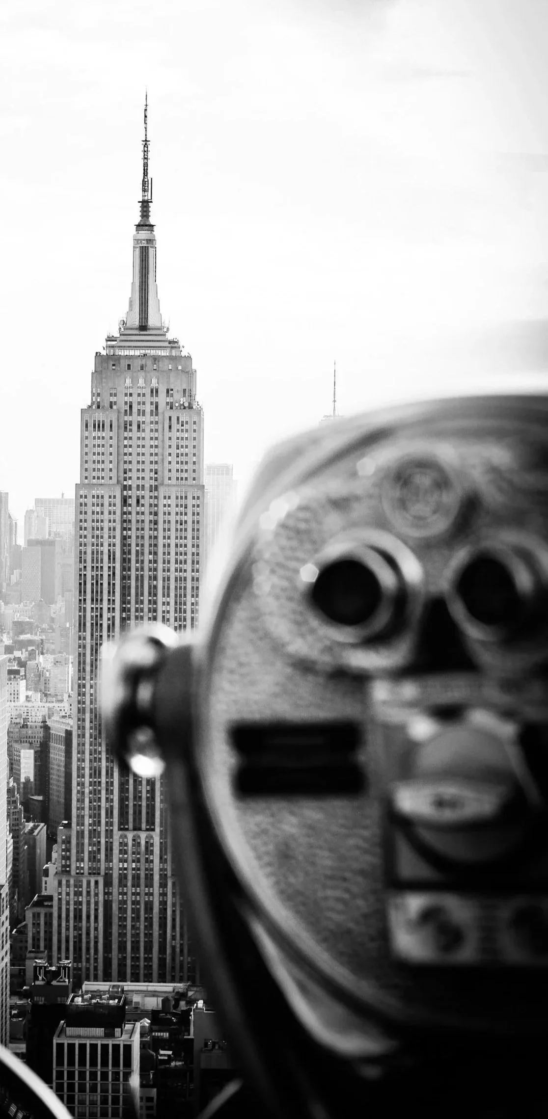 Close-up of a coin-operated binocular viewer with the Empire State Building in focus in the background, on a black and white cityscape