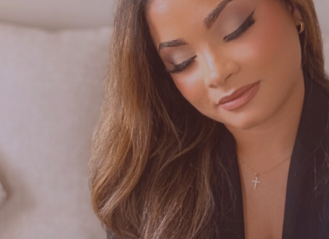 A woman with long, wavy brown hair, wearing makeup with winged eyeliner, light blush, and nude lipstick, with a small cross necklace, closing her eyes and smiling softly.