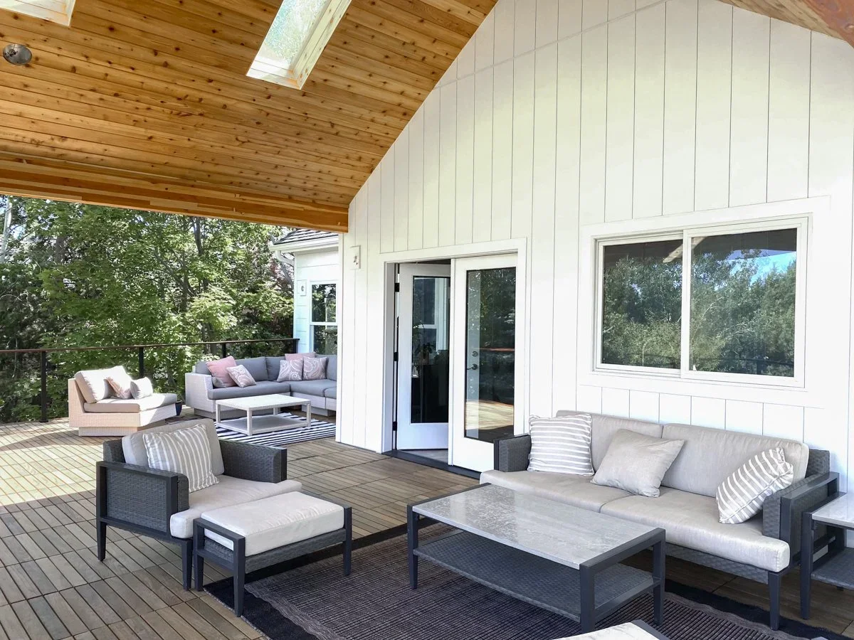 Outdoor patio with seating area, including a sofa, chairs, and a coffee table, under a wooden ceiling with a skylight, overlooking trees and a railing.