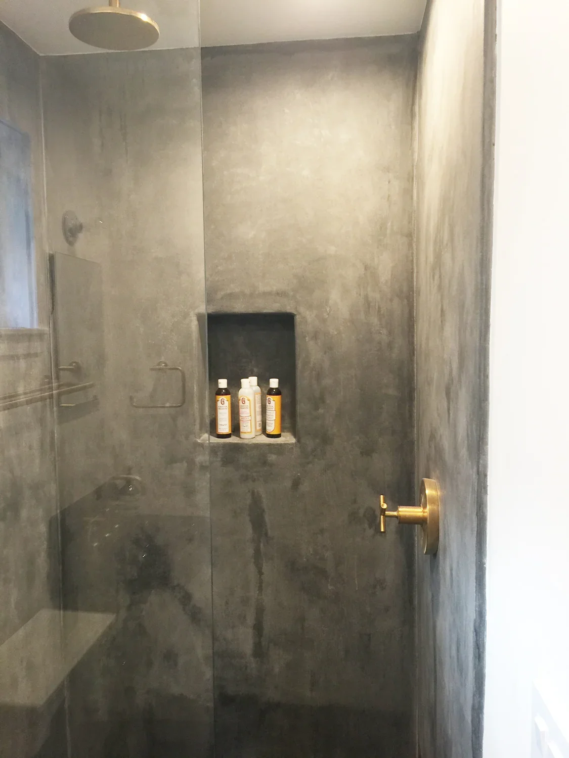 View of a modern shower with gray concrete walls, a built-in wall niche with shampoo and bath products, and gold fixtures including a handle and showerhead.