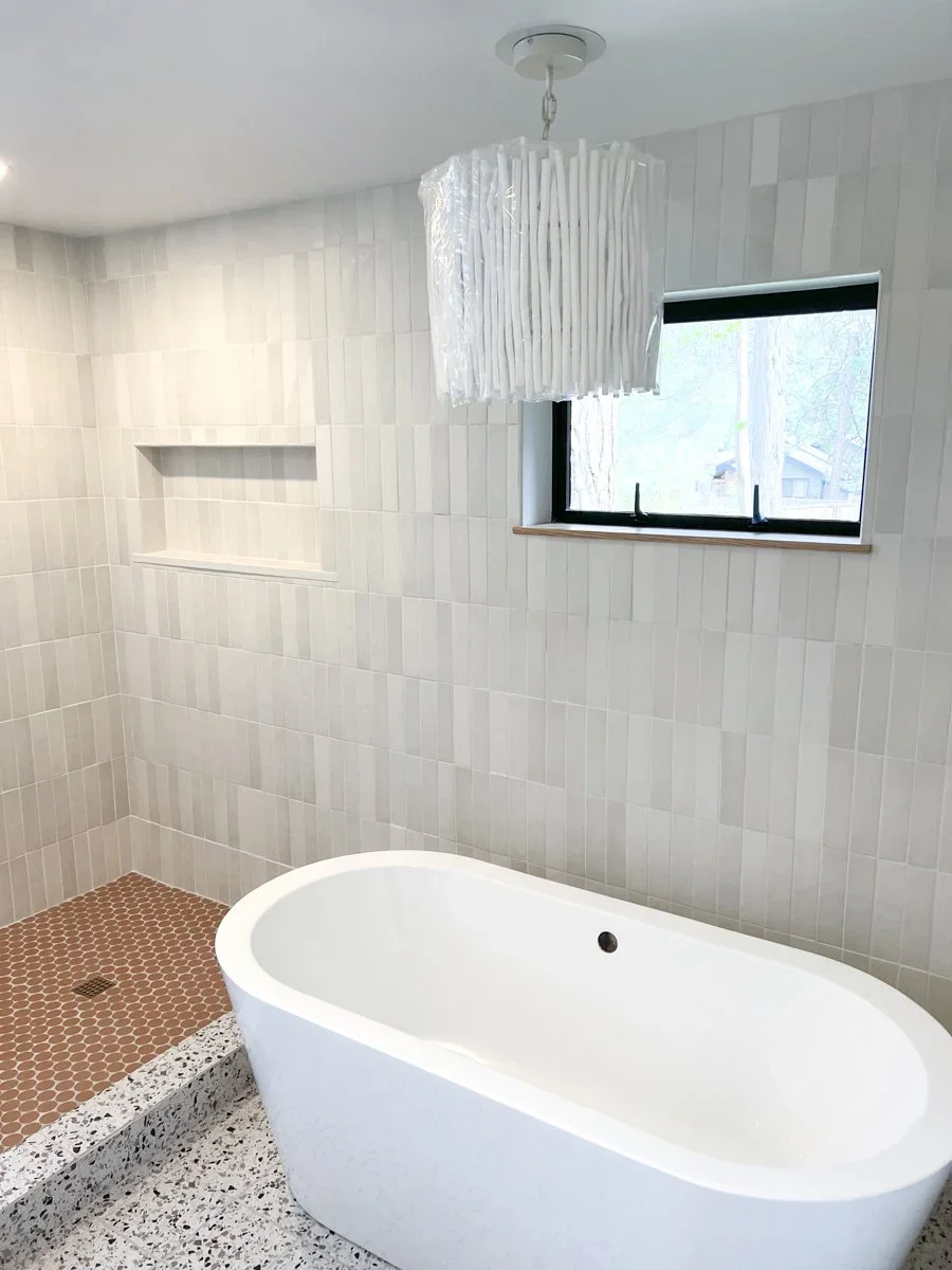 Modern bathroom with white freestanding bathtub, tiled shower area with brown small hexagon tiles, built-in wall niche, window with black frame, and white hanging light fixture.