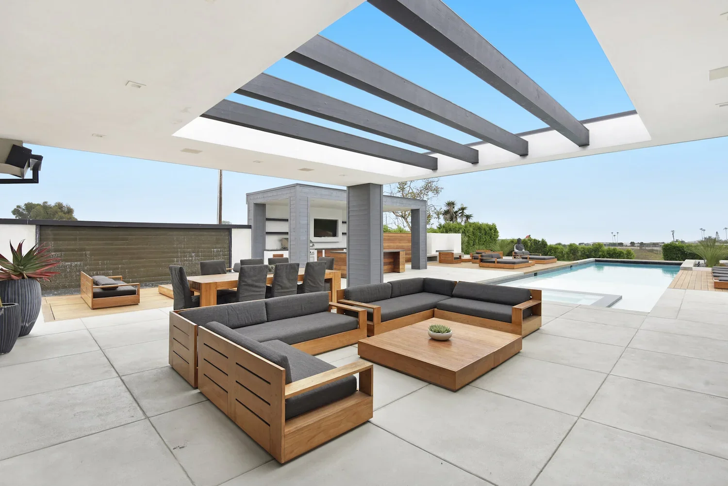 Modern outdoor patio area with gray and wooden furniture, a swimming pool, and a partial roof with skylights, overlooking a clear blue sky.