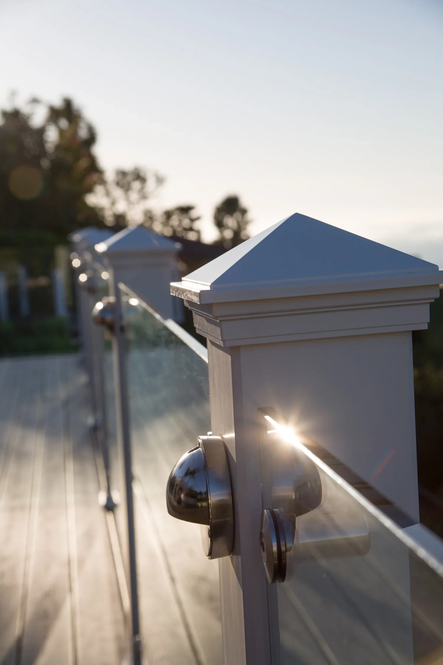 Close-up of a white wooden fence with metal caps on the posts, backlit by the setting sun, with trees in the background under a clear sky.