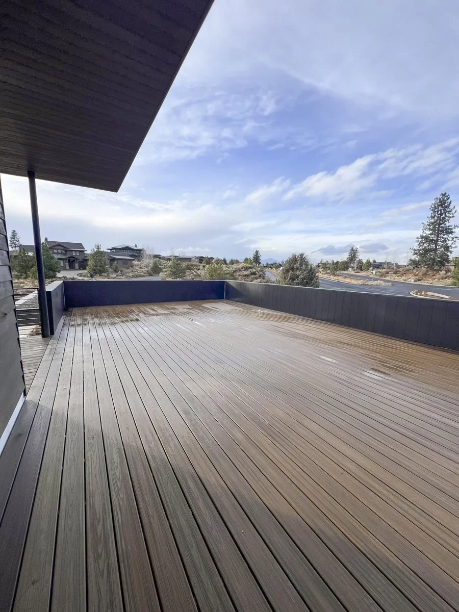 Empty wooden balcony deck with black railing and partial house roof, overlooking a suburban neighborhood with houses, trees, and a partly cloudy sky.