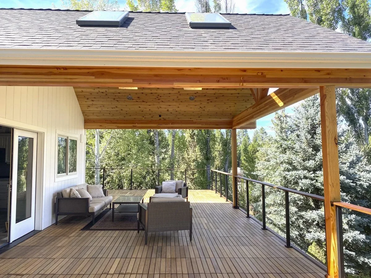 A spacious outdoor balcony with wooden flooring and ceiling, furnished with outdoor seating including a sofa and chairs, overlooking a lush green forest with trees.