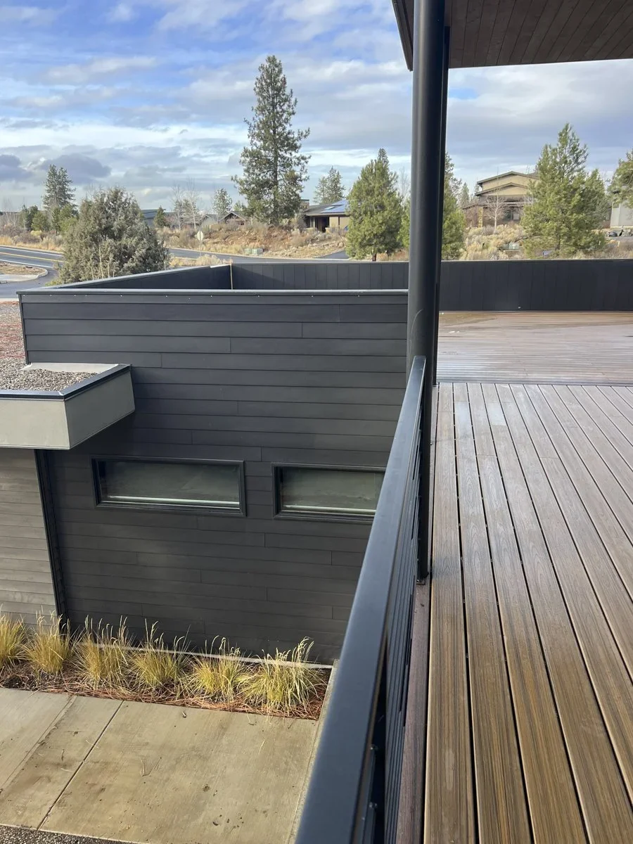Photo of a modern building with dark gray exterior walls and a wooden deck balcony with a black railing, overlooking a landscape with trees and stormy sky.