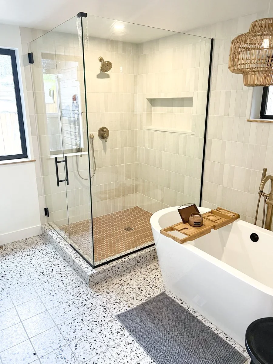 A modern bathroom with a glass-enclosed shower, a bathtub with a tray holding toiletries, and two windows providing natural light. The shower has beige tiles and a built-in shelf, and the bathroom features speckled tile flooring.