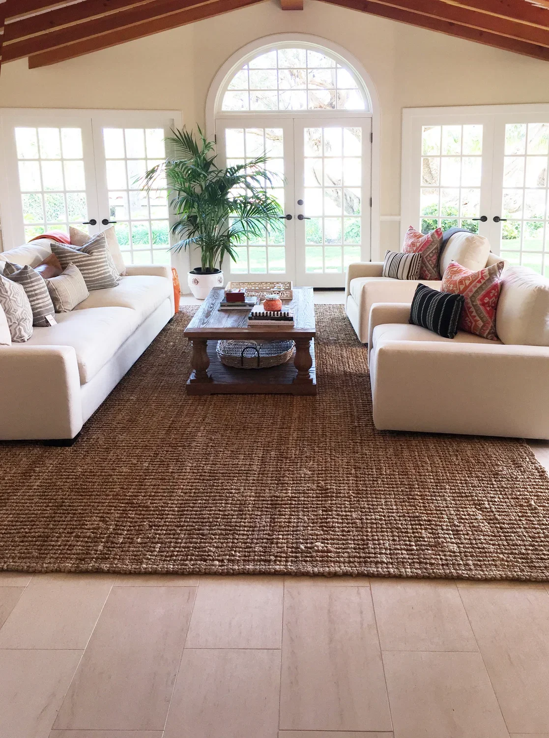 Living room with two white sofas, a large potted plant, wooden coffee table, and large glass doors letting in natural light.