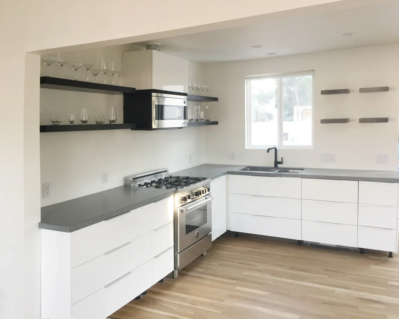 Modern kitchen with white cabinets, gray countertops, a stainless steel stove, black shelves with glassware, and a window above the sink.