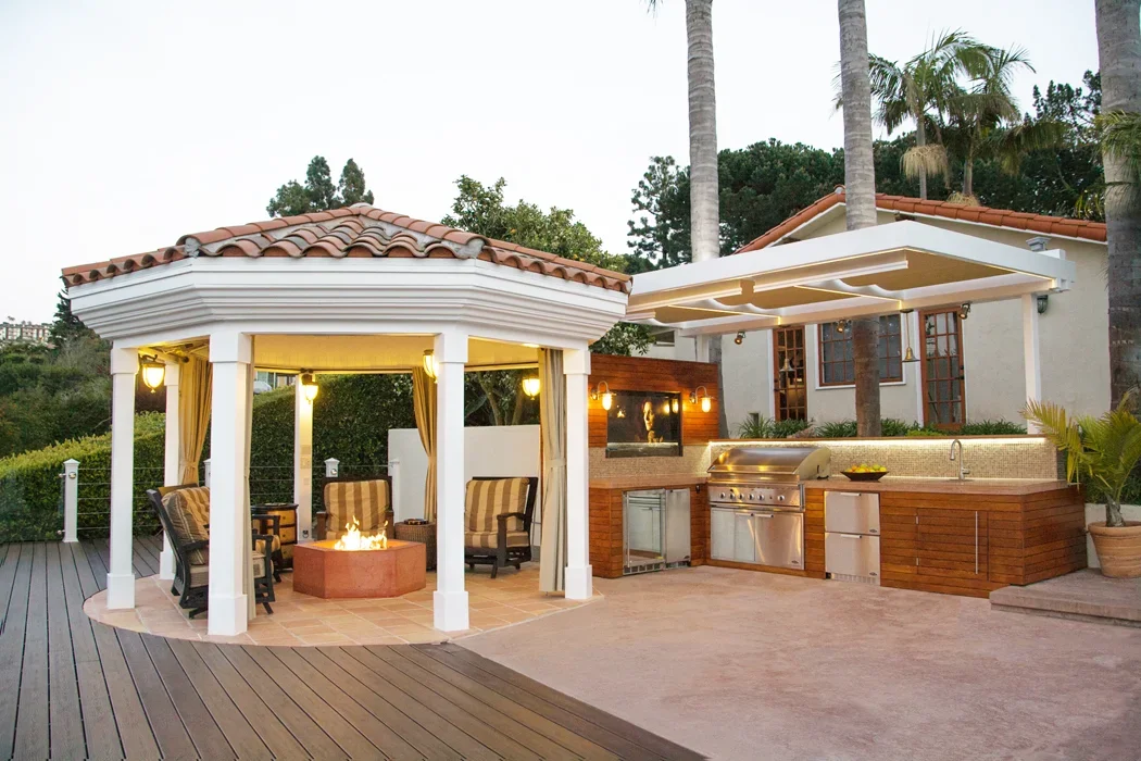 Outdoor patio with a fire pit, seating area, outdoor kitchen with grill and countertop, and a house with a covered porch, surrounded by palm trees and greenery.