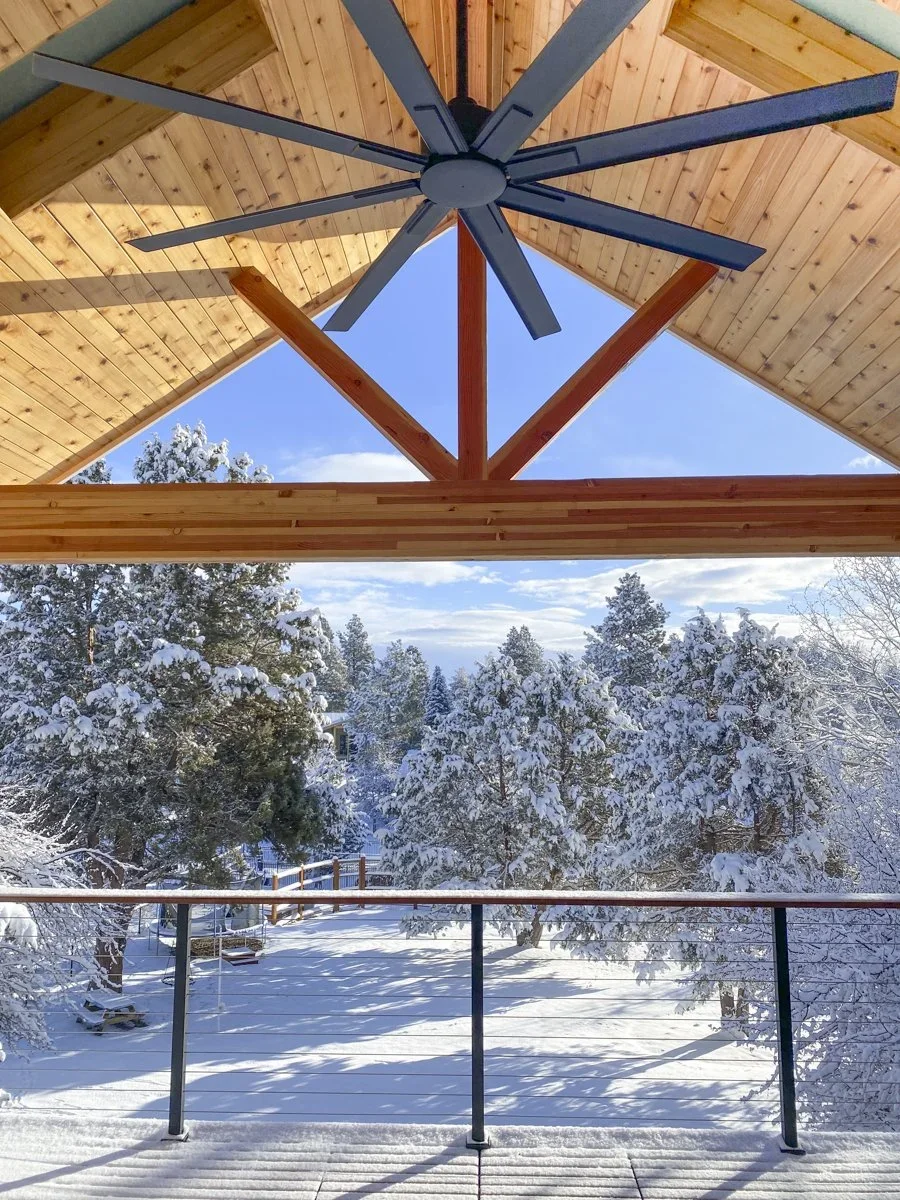 View from a wooden balcony with a black metal railing, overlooking a snow-covered landscape with trees and a blue sky, and a ceiling fan installed under a wooden roof.