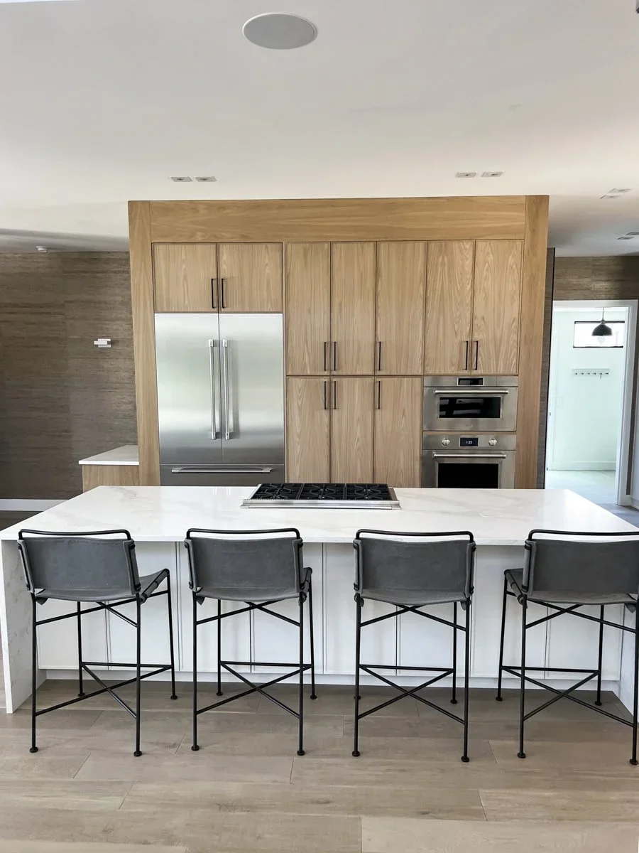 Modern kitchen with white island counter, four black metal and gray chairs, wooden cabinets, stainless steel refrigerator, and built-in oven and microwave.