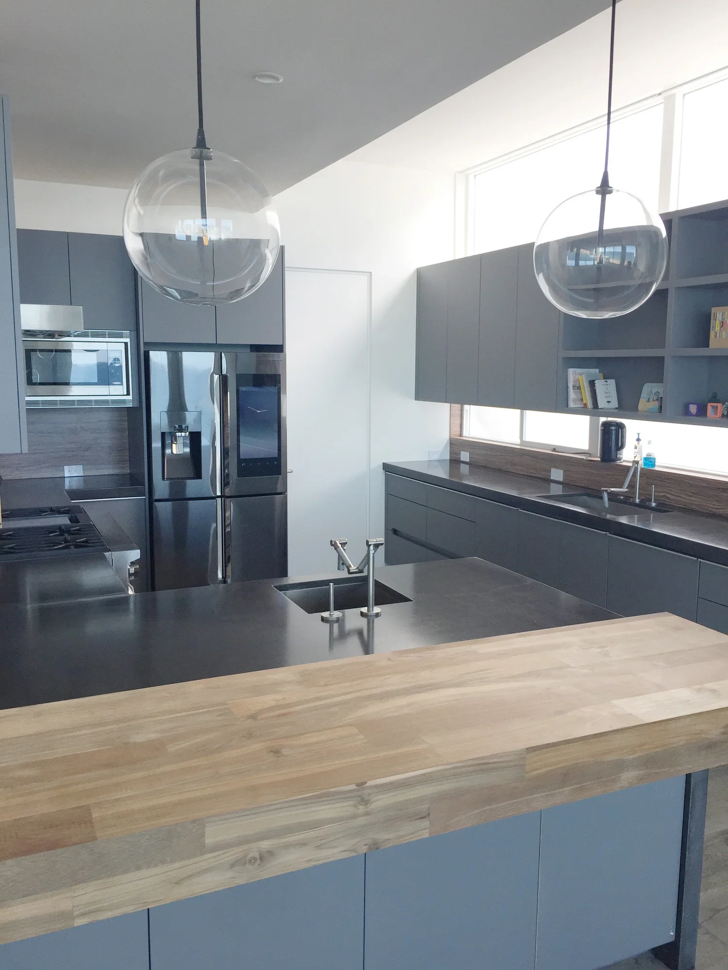 Modern kitchen with gray cabinets, stainless steel refrigerator, built-in microwave, black electric kettle, and a wooden countertop island under two hanging glass pendant lights.