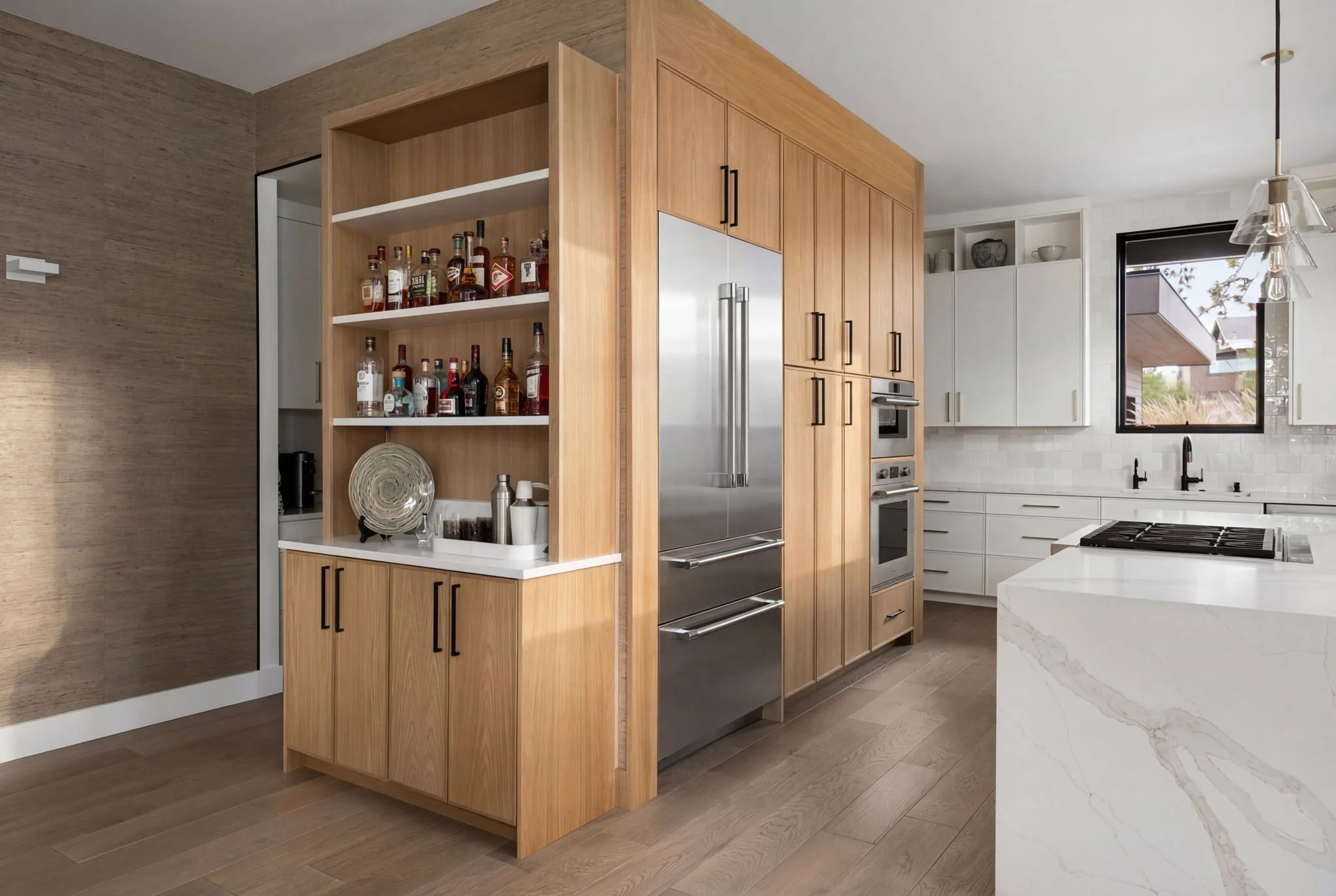Modern kitchen with wooden cabinets, stainless steel appliances, open shelving with liquor bottles, and white countertops.