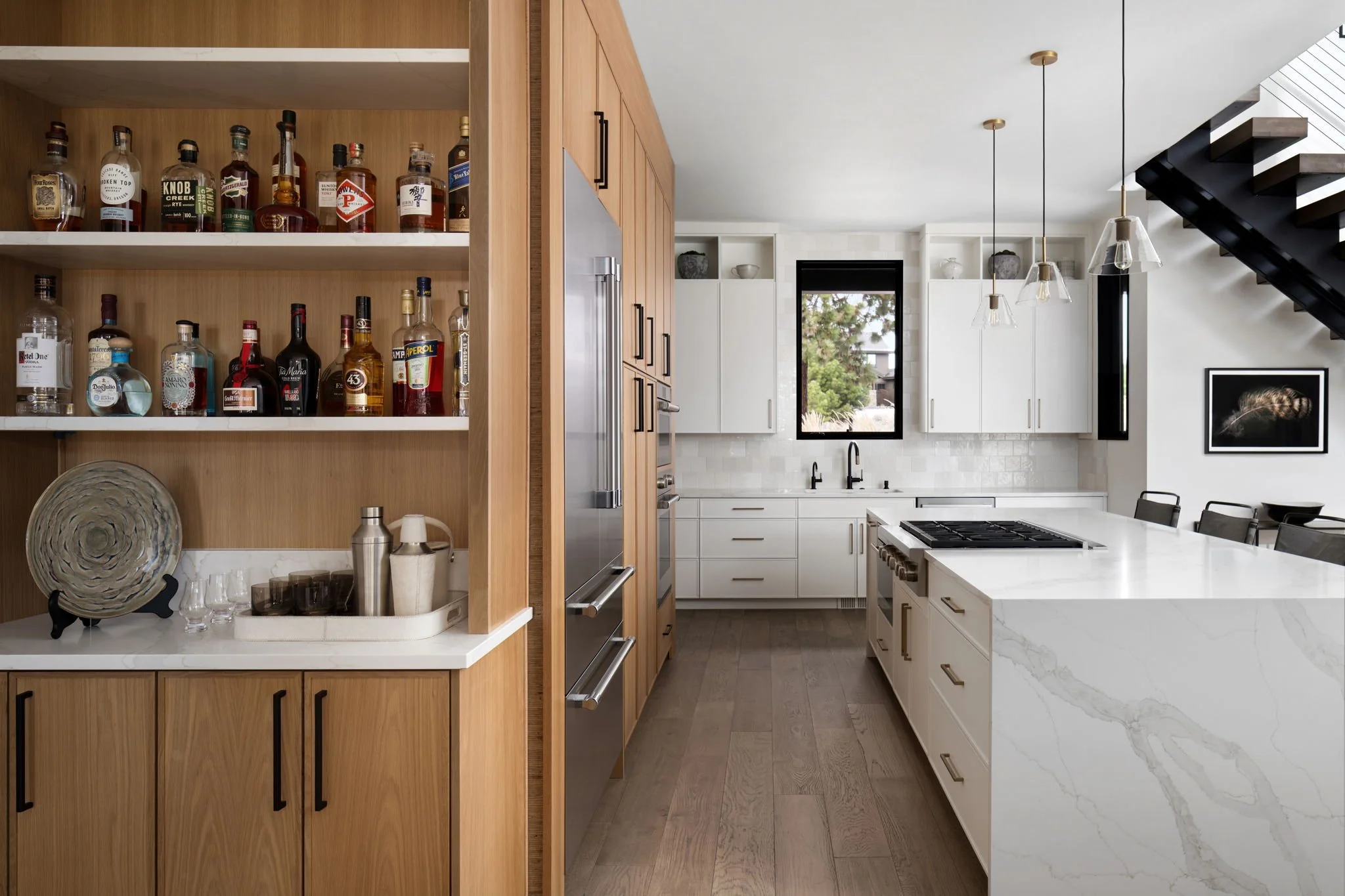 Modern kitchen with white cabinets, a marble island, wooden flooring, and open shelving with liquor bottles.