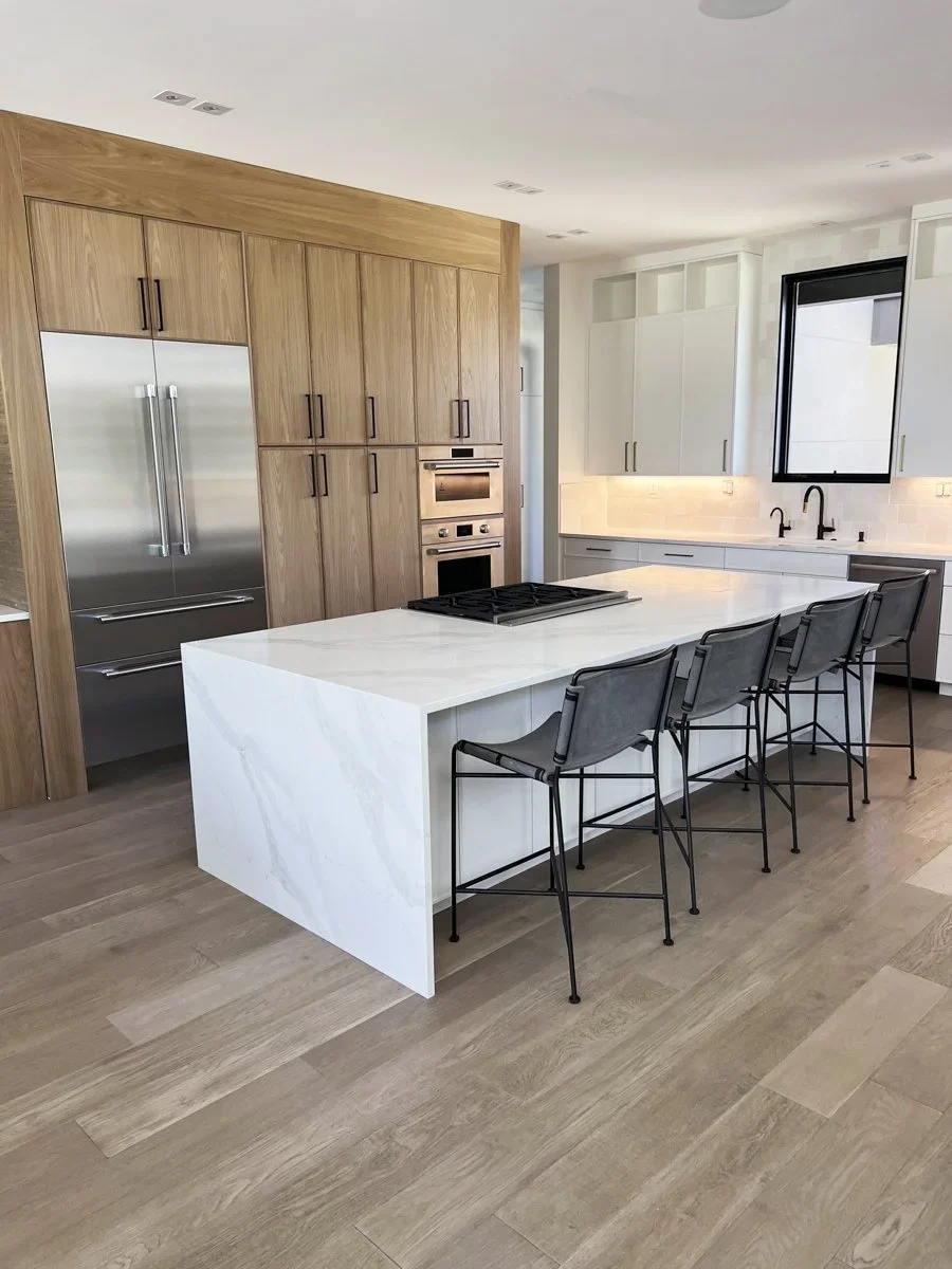 Modern kitchen with wooden cabinets, stainless steel refrigerator, white marble island, and black bar stools.