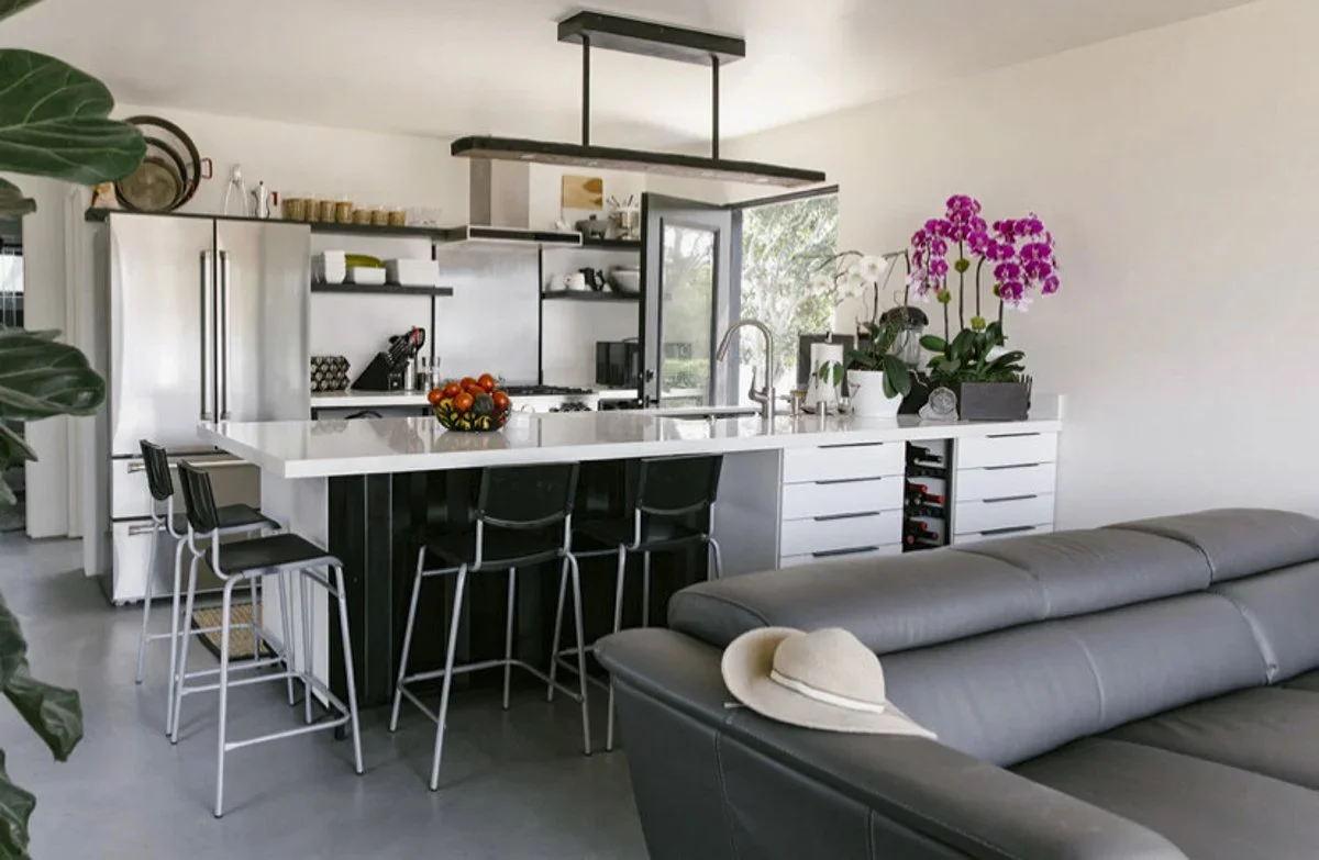 Modern kitchen with white countertops, a black island with four black chairs, and a gray couch with a beige hat resting on the armrest. There is a bowl of tomatoes on the counter, a large pink orchid plant, and an open window with a view of greenery 