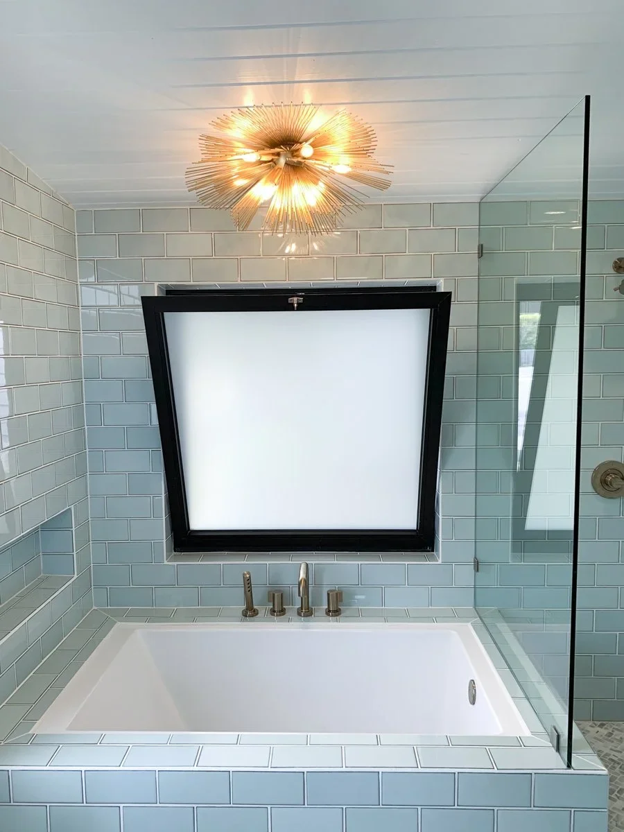 Modern bathroom bathtub with a frosted window above it, white subway tiles, a glass shower wall, and a gold starburst ceiling light fixture.