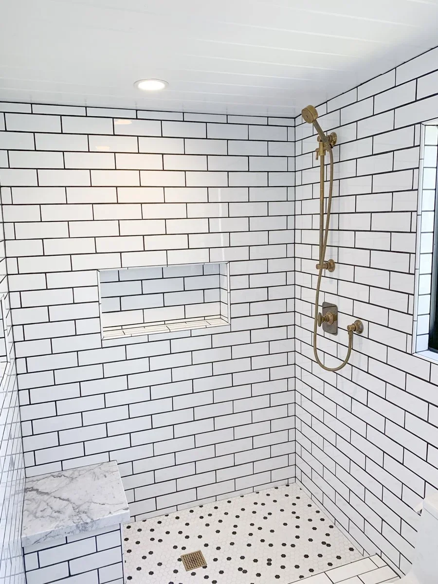 Modern walk-in shower with white subway tile walls, brass fixtures, a built-in niche, marble bench, and small hexagonal tile floor with a drain.