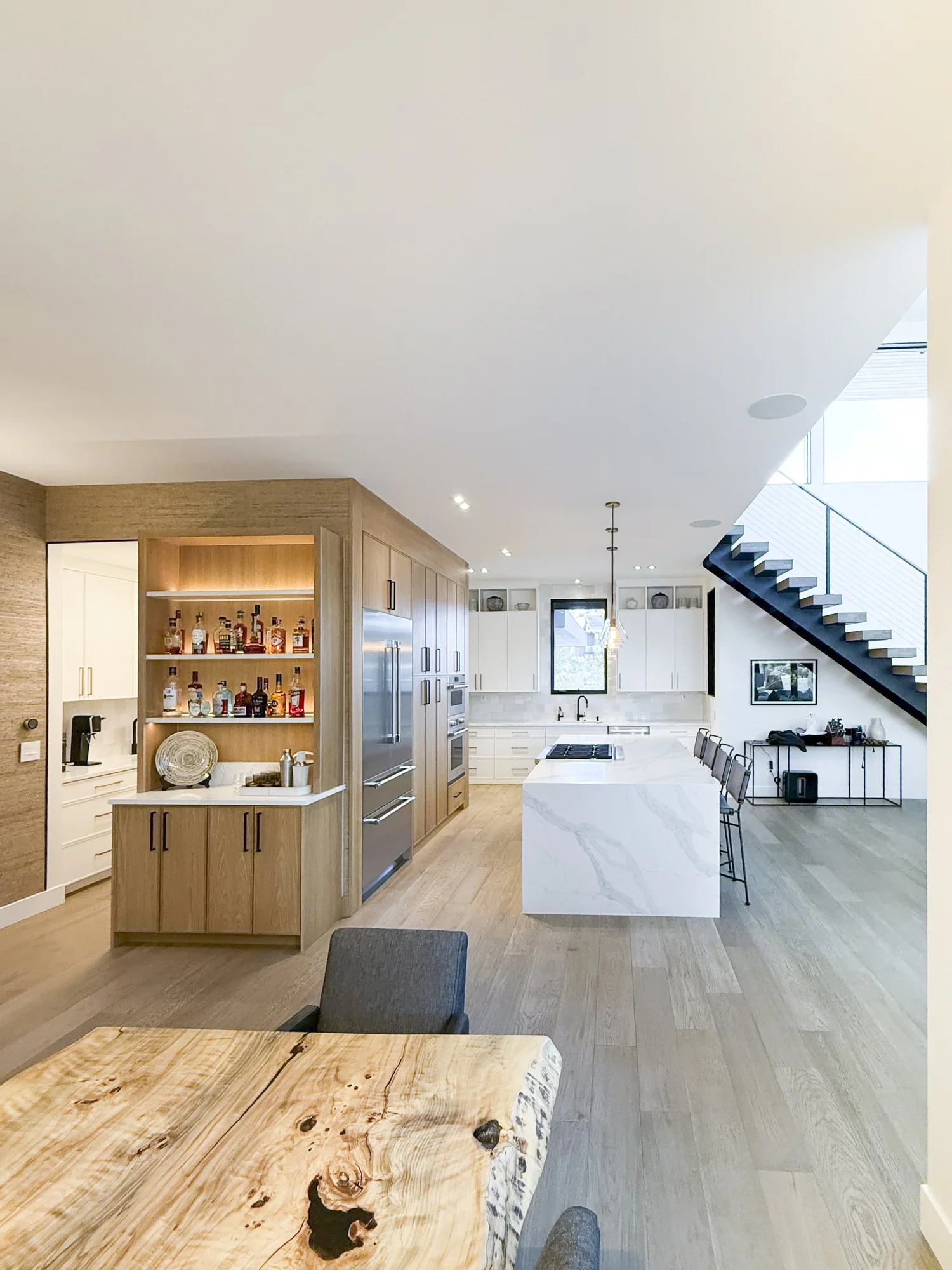 Modern kitchen with white marble island, wooden cabinetry, and black staircase railing, with a wooden dining table in the foreground.
