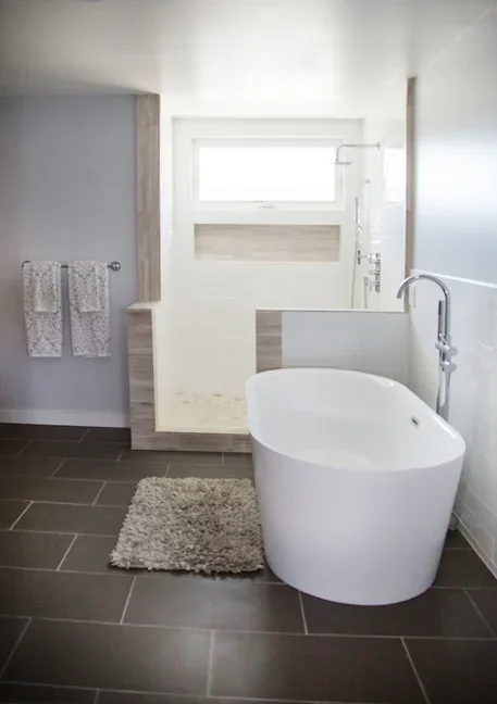 Modern bathroom with freestanding bathtub, gray floor tiles, and a window above the bathtub.