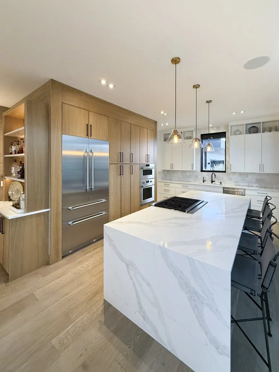 Modern kitchen with white marble island counter, wooden cabinetry, stainless steel refrigerator, built-in oven, black faucet, minimalist decor, and pendant lights.