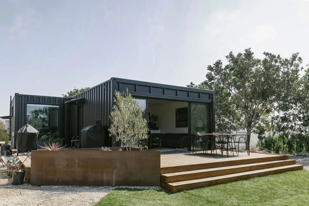 Modern tiny house with a black exterior, sliding glass door, outdoor dining area, and wooden steps leading to a grassy yard, surrounded by trees.