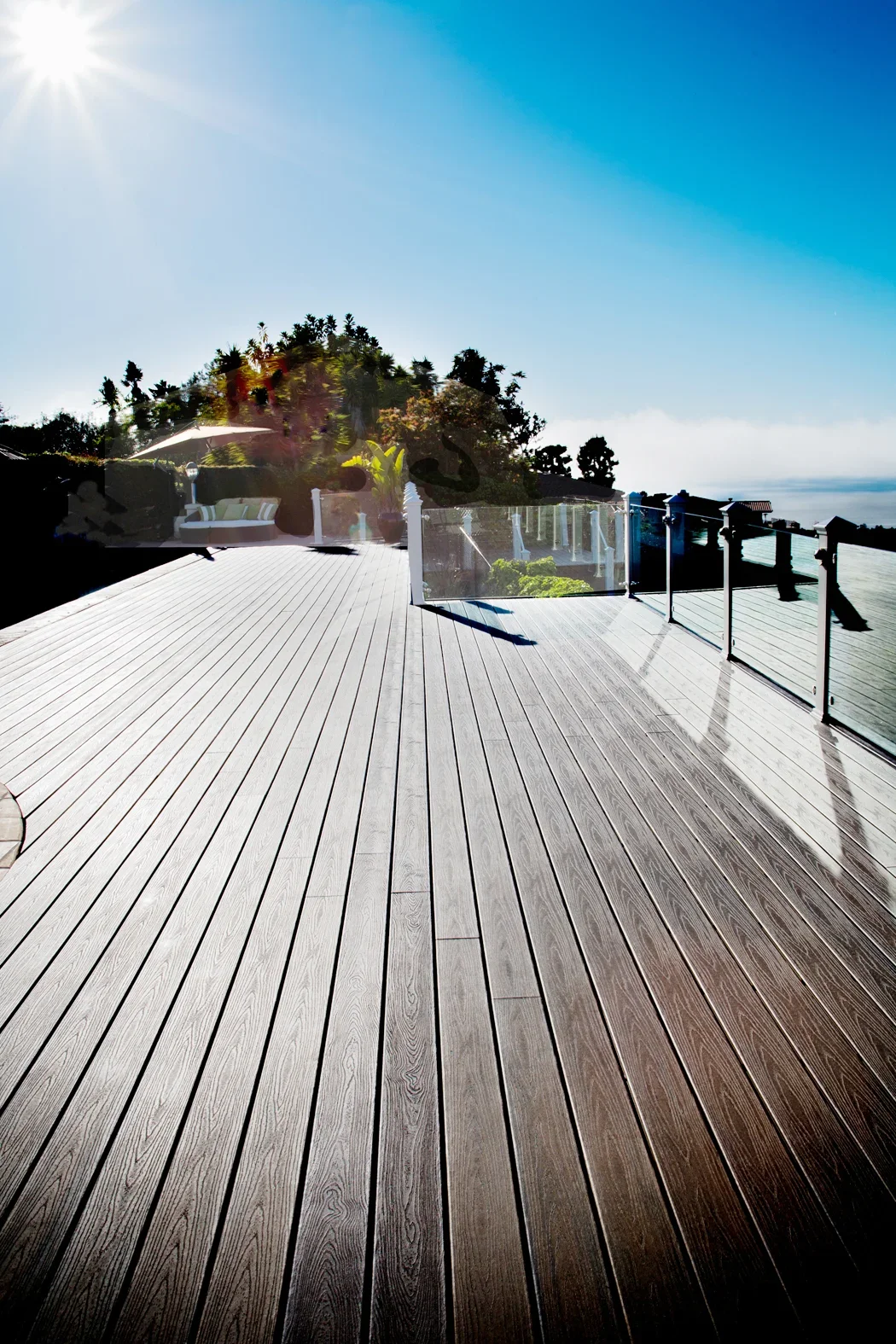 A spacious outdoor deck with wood flooring, glass railing, greenery, outdoor furniture, and a bright blue sky with sunlight.