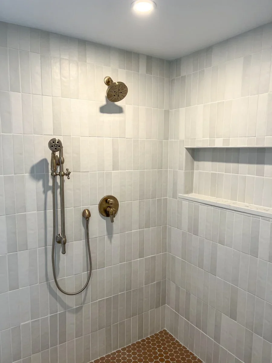 Shower area with white tiled walls, a brass showerhead, and a shower control handle, featuring a built-in wall niche for storage, and a small recessed ceiling light.