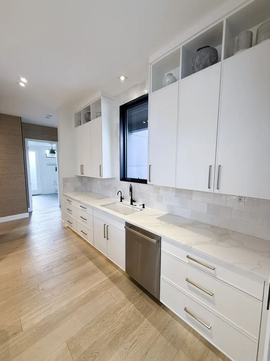Modern kitchen with white cabinets, marble countertops, black faucet, undermount sink, and stainless steel dishwasher. Light wood flooring and decorative vases on upper shelves. Large window above sink, bright and minimalistic decor.