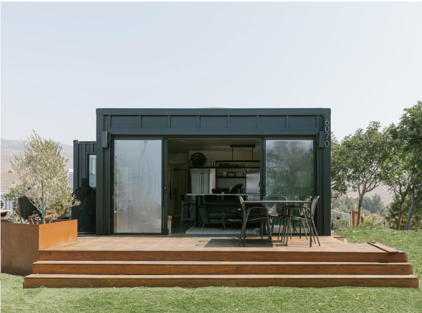 Modern tiny house with a wood deck, outdoor dining table, and chairs, large glass sliding doors revealing a kitchen and living area inside, surrounded by trees and greenery.