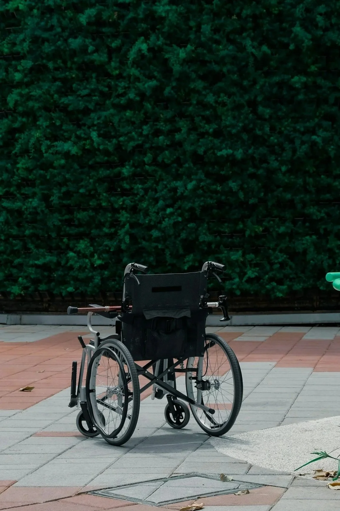 A wheelchair is parked on a sidewalk in front of a tall green hedge, with a traffic light to the right.