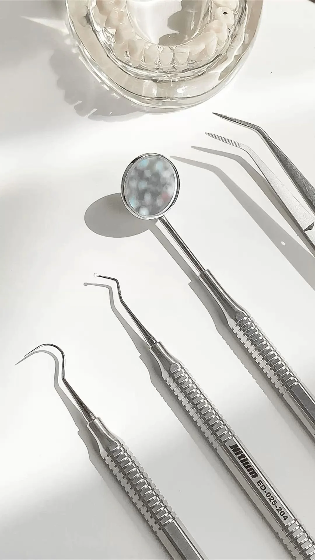 Close-up view of dental tools, including a dental mirror and probes, arranged on a white surface with a glass container filled with small white objects in the background.