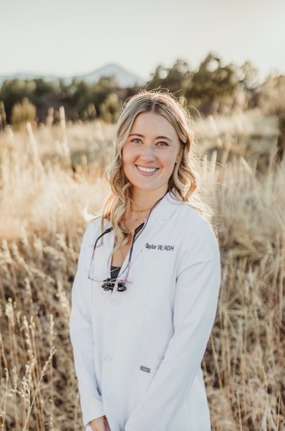 A young woman in a white medical coat standing outdoors in a field of tall, dry grass with mountains and trees in the background, smiling at the camera.