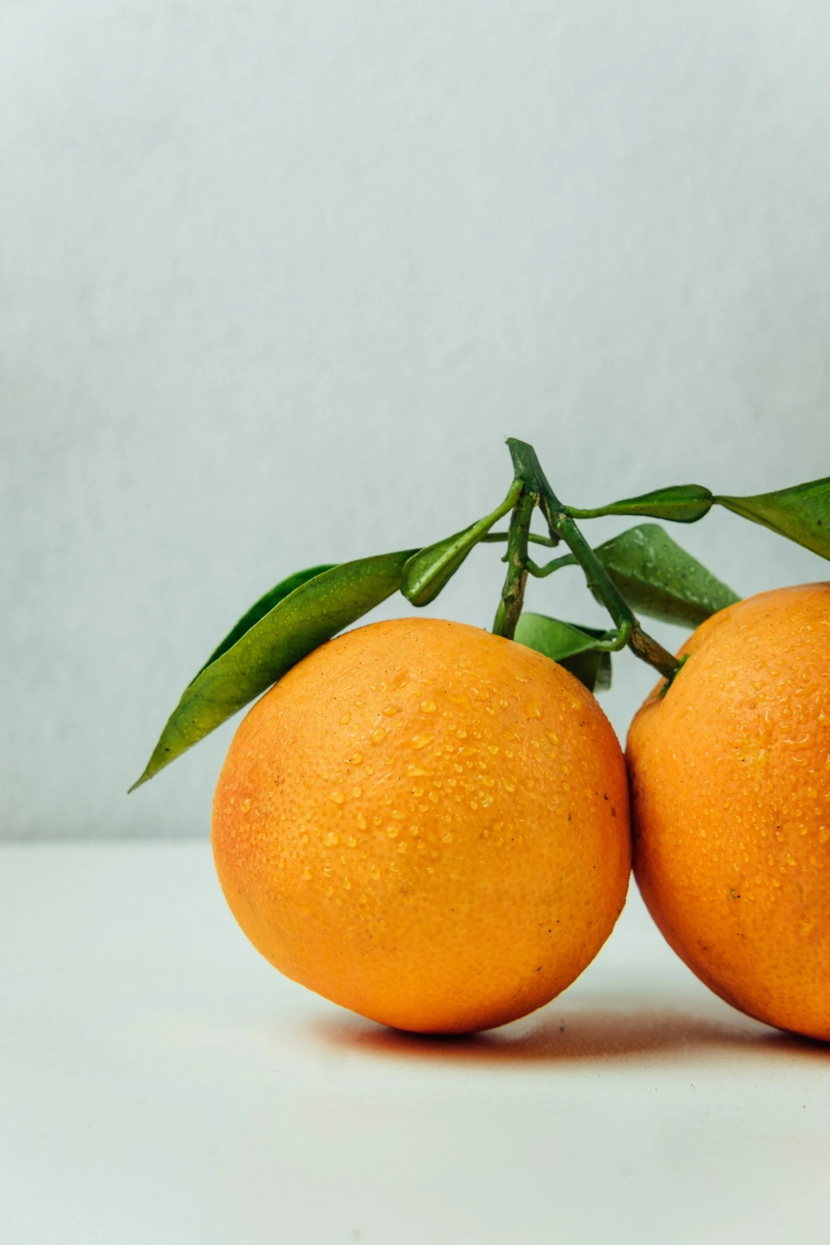 Two fresh oranges with green leaves and water droplets on their surfaces, placed on a white surface against a light background.