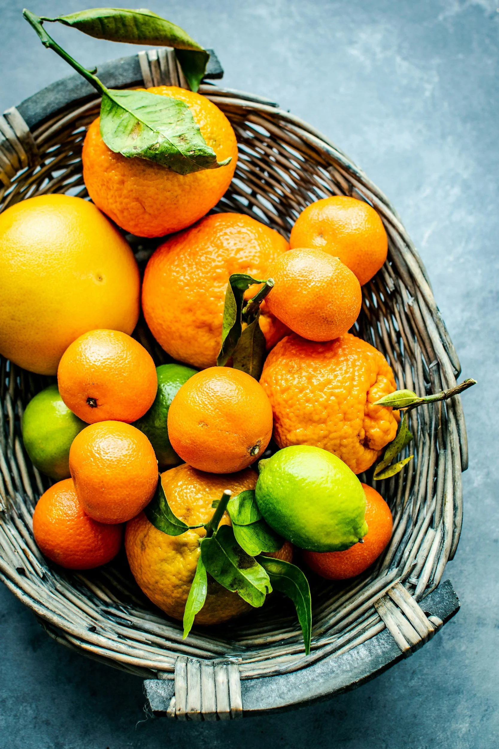 A wicker basket filled with various citrus fruits including oranges, lemons, limes, and tangerines, some with green leaves attached, on a dark gray surface.