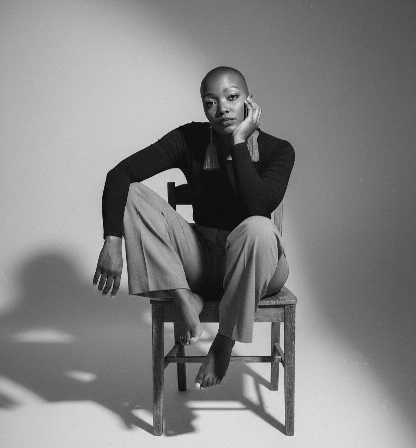 Black and white photo of a woman sitting on a chair with feet on the chair's seat, wearing a long sleeve top, wide pants, and tassel earrings, posed with her chin resting on her hand.