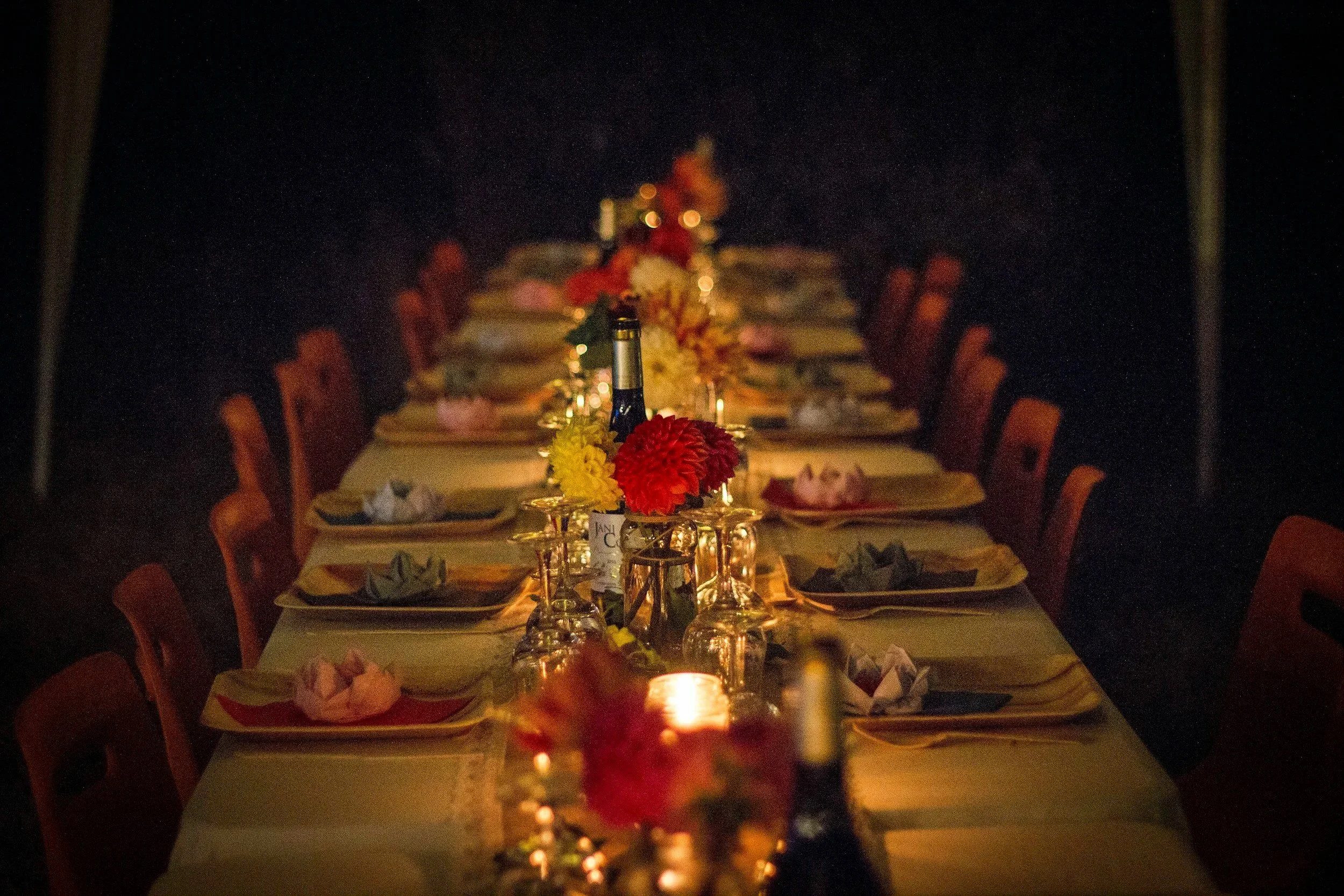 A long dining table decorated with flowers, glasses, and place settings, dimly lit with candles, set for an event or dinner gathering at night.