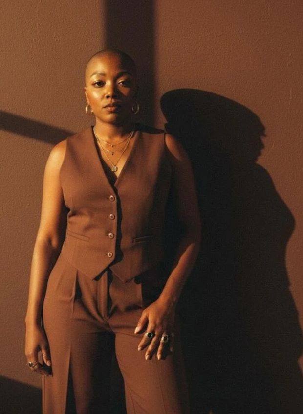 A woman with a shaved head standing against a brown wall with dramatic shadow, wearing a brown sleeveless vest and matching pants, accessorized with gold jewelry and rings.
