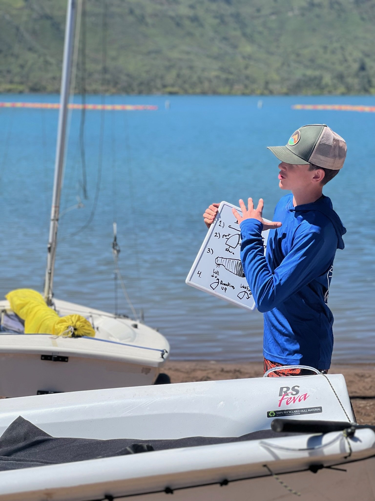 A boy standing on a dock by a lake, wearing a blue jacket and a cap, holding a whiteboard with sketches and notes related to sailing.