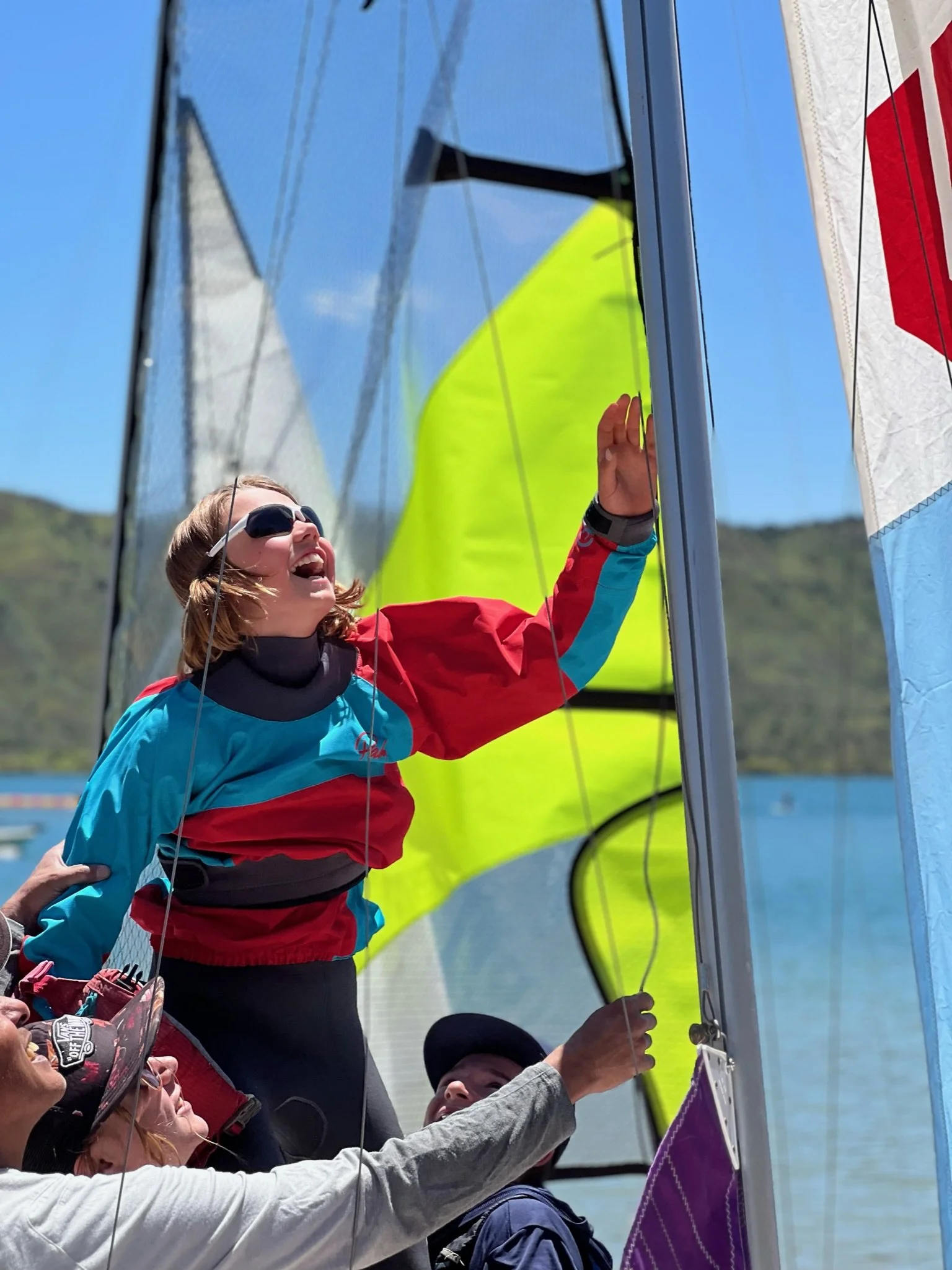 A woman in sunglasses and a colorful windbreaker smiling and pointing at a sail, surrounded by other people on a sailboat, with water and a hilly landscape in the background.