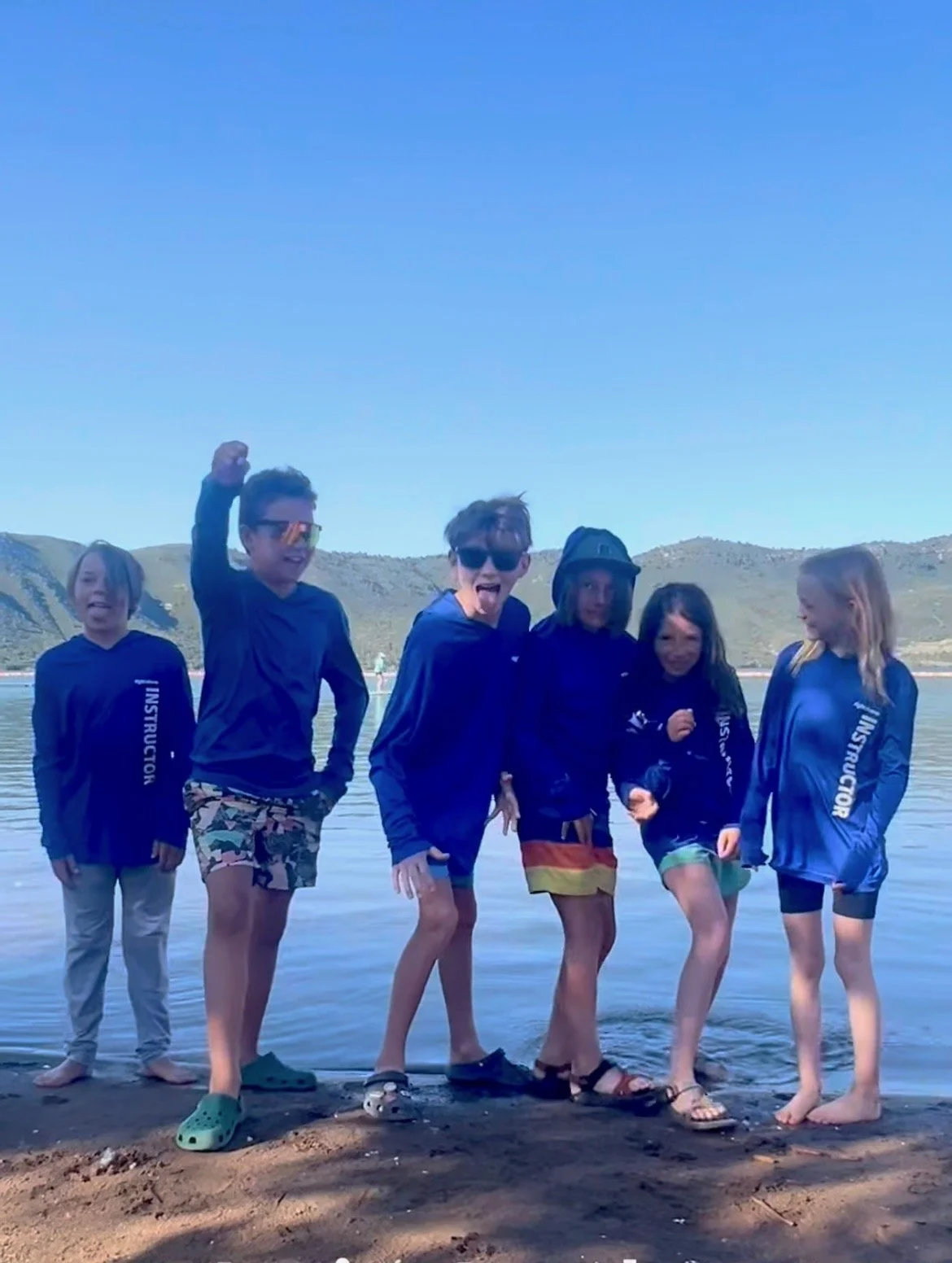 Six children on a beach near a body of water, some wearing matching blue jackets with 'INSTRUCTOR' written on the sleeves, smiling and posing for the photo with hills in the background.