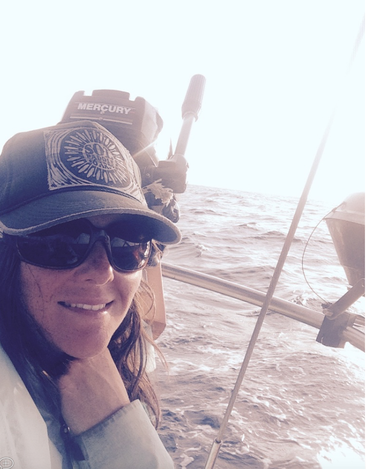 A woman on a boat taking a selfie with the ocean in the background. She is wearing sunglasses, a cap, and smiling.