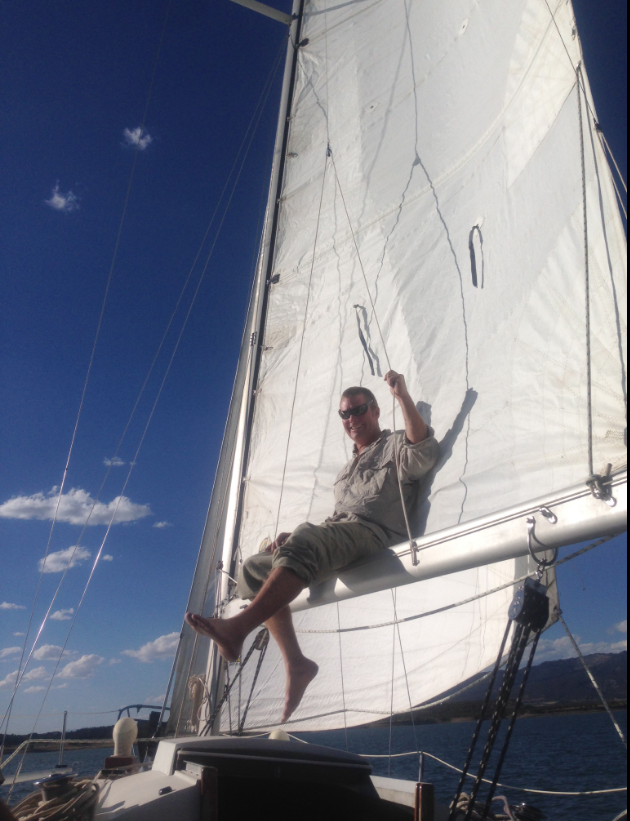A man sitting on the sailboat's bow, smiling and holding onto the sail's ropes, with the blue sky and water in the background.