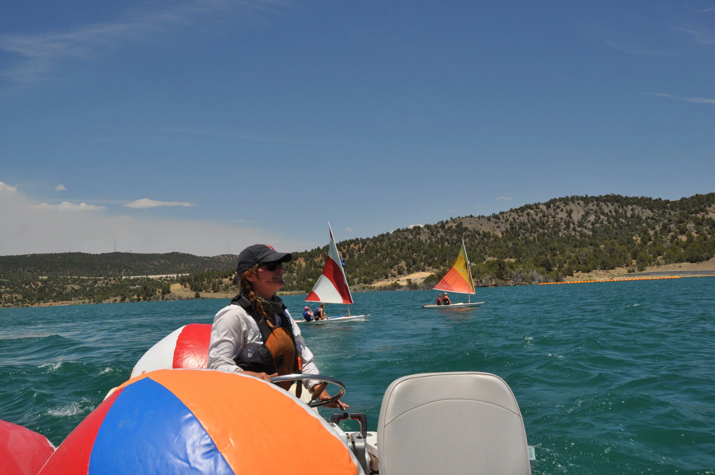 Children sailing during summer camp with a person in the foreground on a boat, with hills and a clear blue sky in the background.