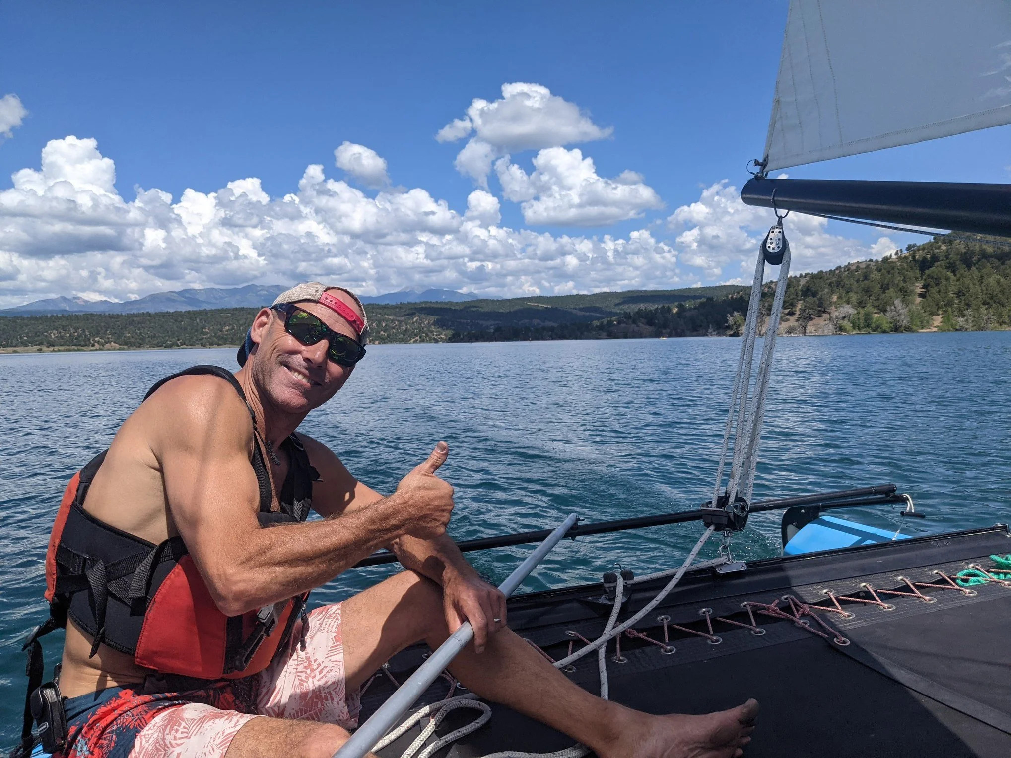 Man on a sailboat giving a thumbs up, wearing sunglasses, a sandy cap, and swim trunks, on a lake with trees and mountains in the background.