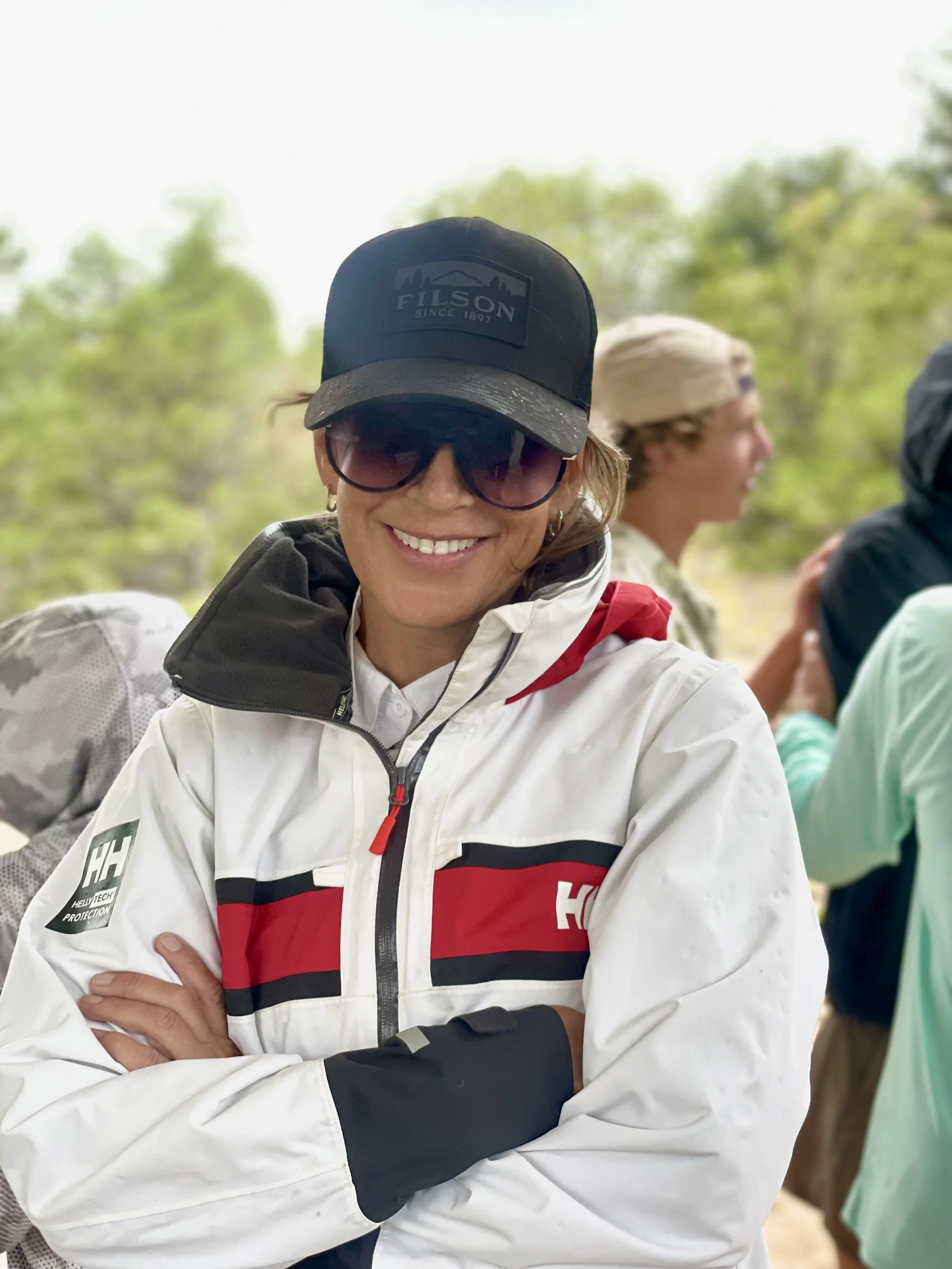 A woman wearing a black baseball cap, sunglasses, and a white Helly Hansen jacket with red and black stripes, smiling with her arms crossed outdoors with blurred trees in the background.