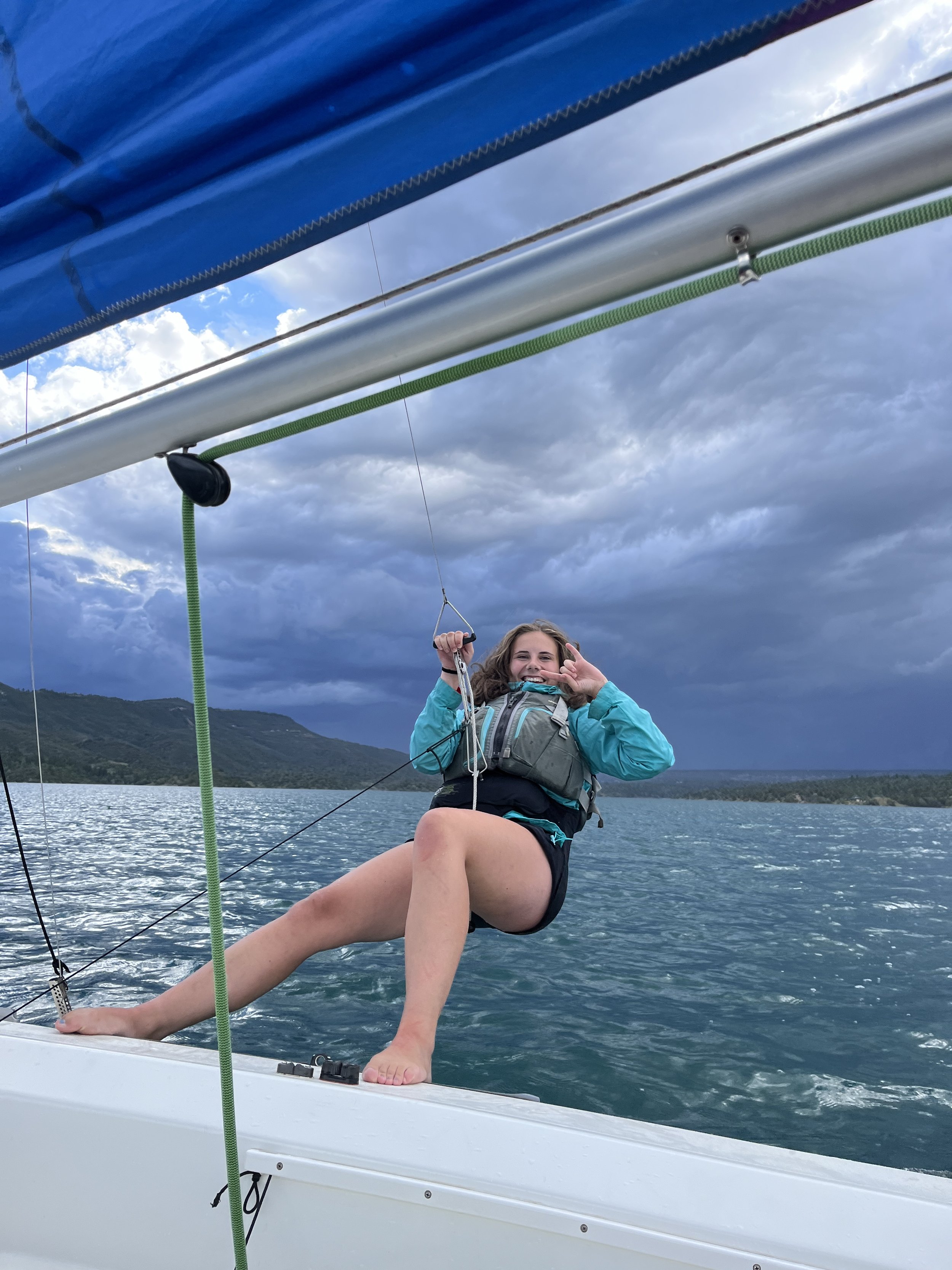 Woman in a life jacket smiling and making a peace sign while sitting on the edge of a sailboat on a lake with mountains and a cloudy sky in the background.