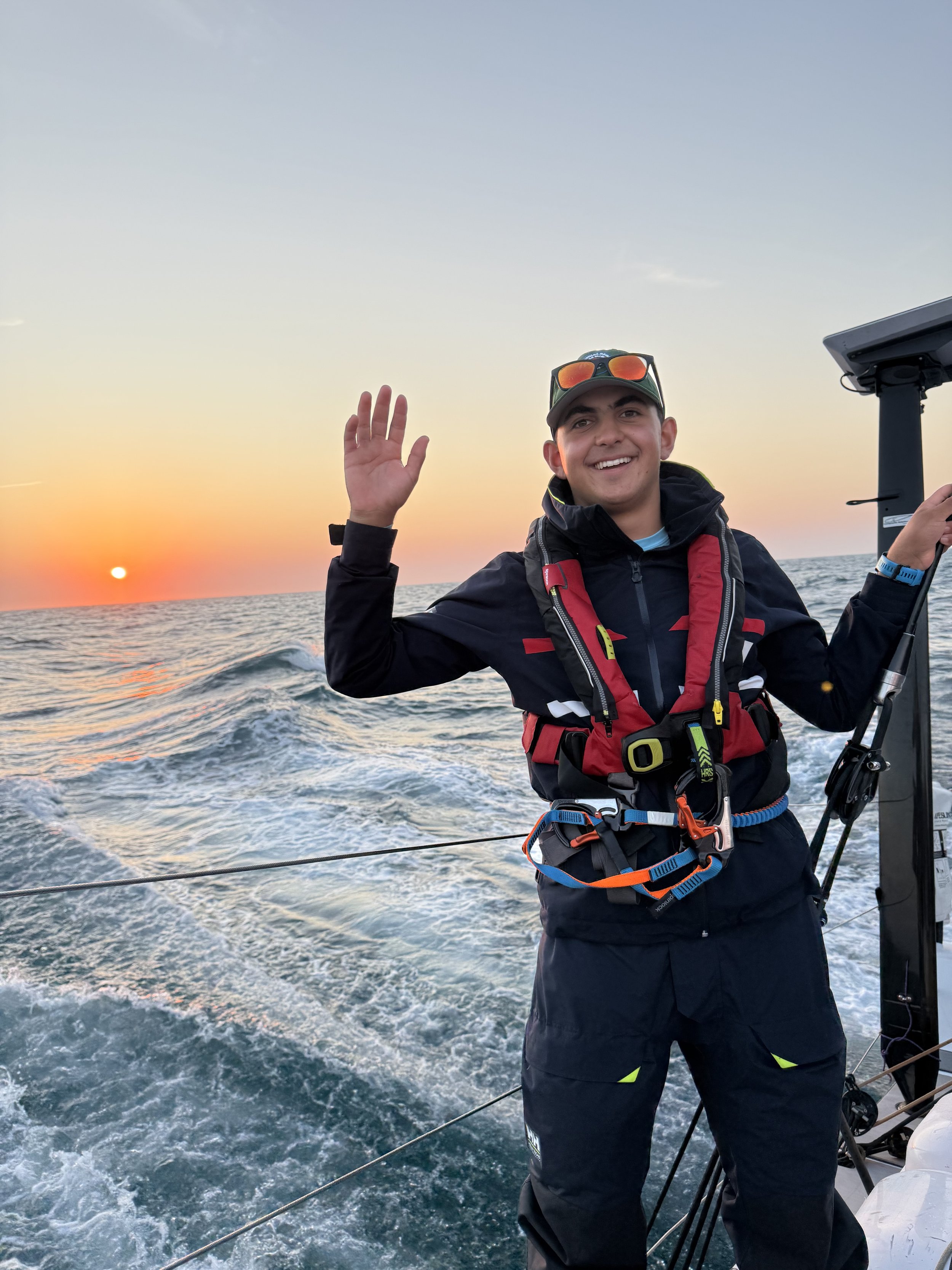 Young man smiling and waving on a boat at sunset, wearing sailing gear and sunglasses on his cap, with the open ocean and a setting sun in the background.